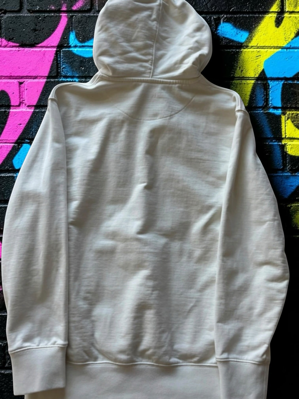 Back view of a plain white Aurora Fashion Palace Skateboards cotton pullover hoodie against a graffiti brick wall.